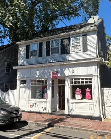 White colonial-style storefronts on brick street with parked car and tree shade, clear blue sky