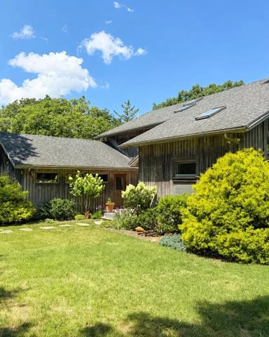 Modern barn-style house with dark wood siding, surrounded by mature trees and landscaped shrubs on a grassy lawn.