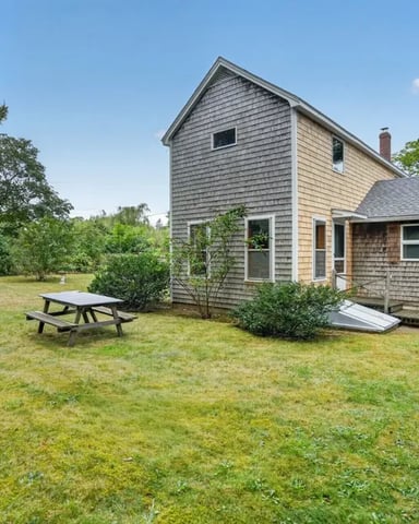 Shingled cottage with deck and picnic table on grassy lawn surrounded by trees