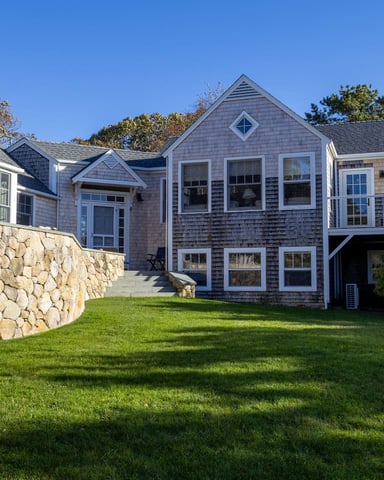 Blue and gray shingled coastal house with stone retaining wall and manicured lawn