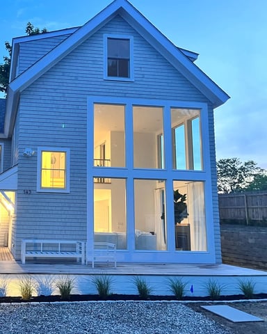 Blue modern cottage with large glass doors, white deck, and landscaped front yard at dusk