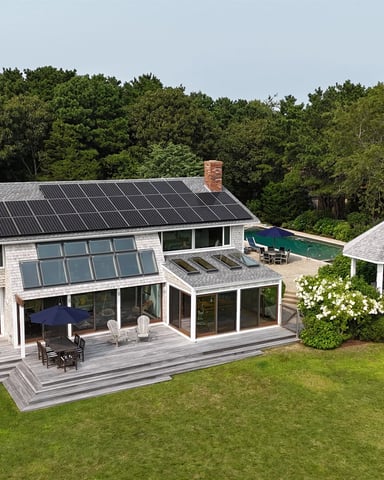 Modern shingle-clad house with solar panels, wraparound deck, pool, and gazebo surrounded by trees