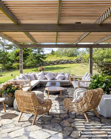Covered patio with wooden pergola, woven chairs, sectional sofa, and stone fireplace overlooking manicured lawn and gardens