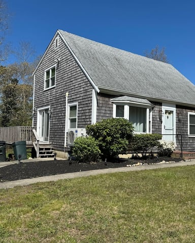 Gray shingled cottage with white trim, front porch, and landscaped yard under clear blue sky