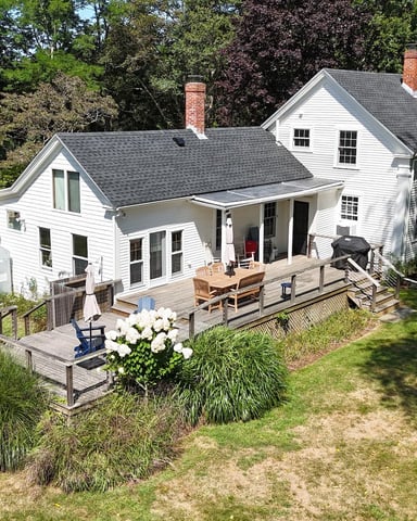 White farmhouse with gray roof, brick chimneys, and wraparound deck surrounded by mature trees and landscaping.