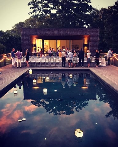 Evening outdoor event with guests gathered on wooden deck beside illuminated pool, modern dark-shingled structure with large windows, string lights and