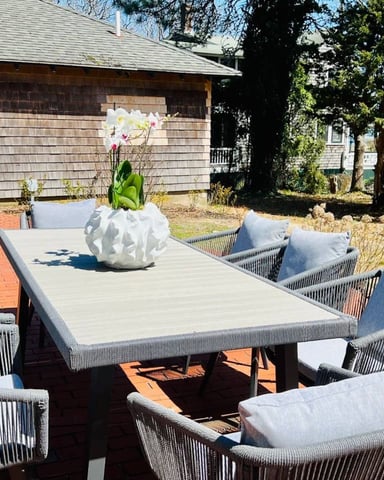 Outdoor patio with gray woven chairs, rectangular table, white orchid centerpiece, and brick pavers beside shingled cottage