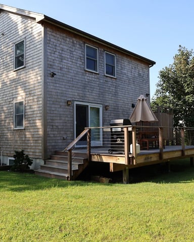 Modern gray shingle house with wooden deck, patio umbrella, and small shed surrounded by green lawn and trees.
