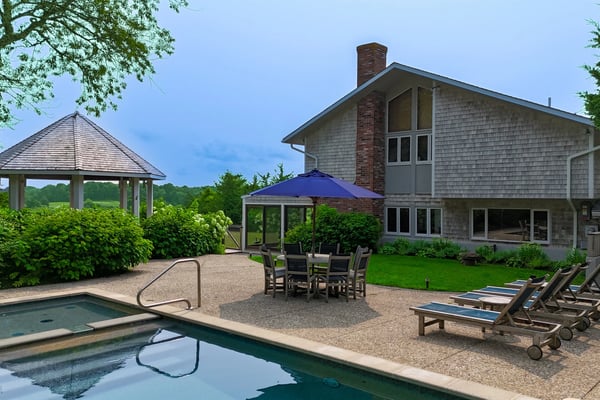 Modern shingle-clad house with pool, hot tub, dining area under blue umbrella, gazebo, and lounge chairs overlooking green lawn and distant fields.