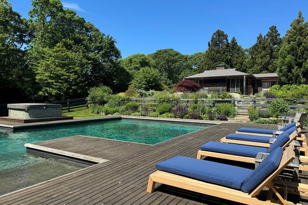Wooden deck with blue lounge chairs overlooking turquoise pool, modern house and manicured gardens surrounded by trees