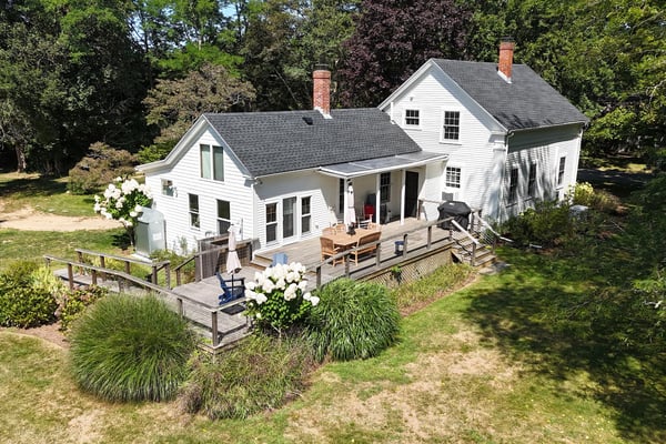White farmhouse with gray roof, brick chimneys, and wraparound deck surrounded by mature trees and landscaping.