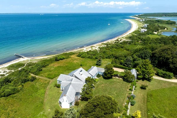 Aerial view of waterfront home on grassy bluff overlooking curved sandy beach and blue ocean