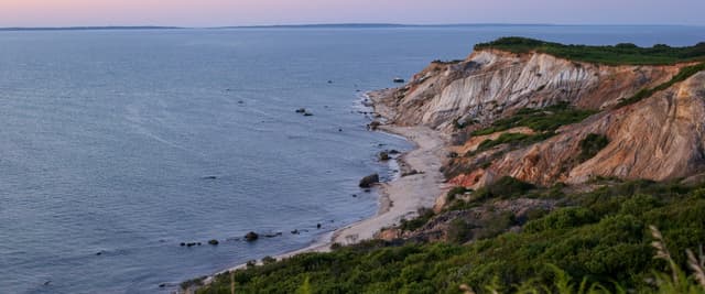 Aerial view of coastal town at sunset with sailboats in harbor, lighthouse on sandy point, and waterfront homes