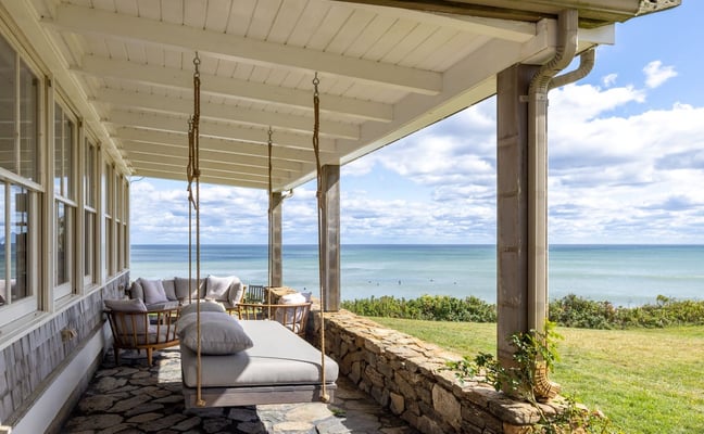 Covered porch with rope swing bed and ocean view, stone wall, wooden columns, and coastal landscape