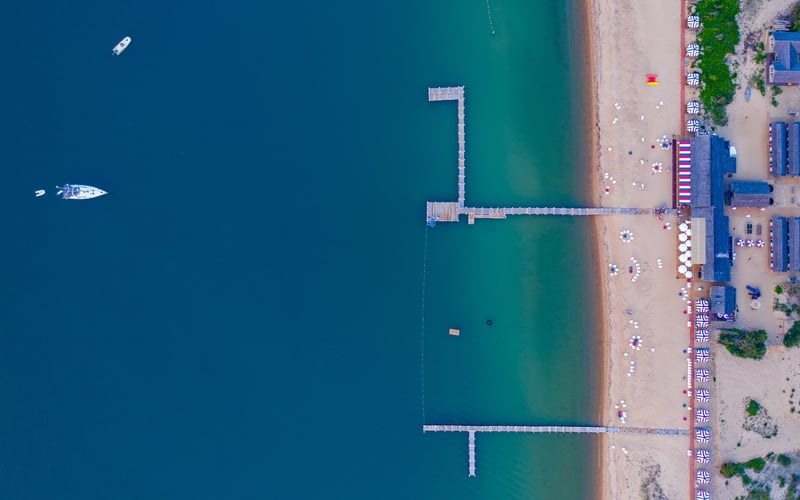 Aerial view of beach with wooden pier, turquoise water, boats, and beachgoers with umbrellas